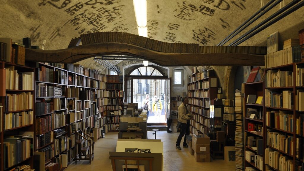 Es ist ein Buchladen. Links und rechts ist ein großes Regal voller Bücher. In der Mitte des Raumes ein Lesetisch.